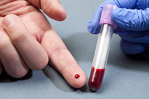 A finger with drop of blood next to a vial of blood