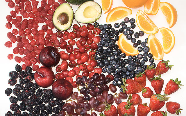 Black plums, blackberries, raspberries, strawberries, sweet cherries, avocado, navel orange, and red grapes on white background.