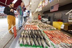 Two male shoppers looking over a selection of fresh fish in a seafood market display case.