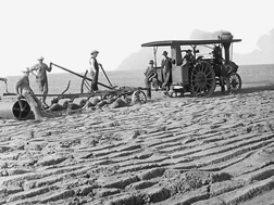 Several men congregate by a tractor-pulled one-way production plow in a partially tilled field.